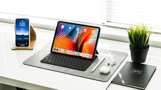 Tablet with keyboard, smartphone, and notebook arranged beside a plant on a bright desk.