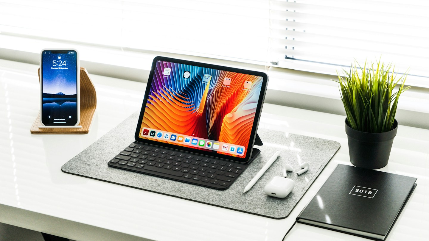 Tablet with keyboard, smartphone, and notebook arranged beside a plant on a bright desk.