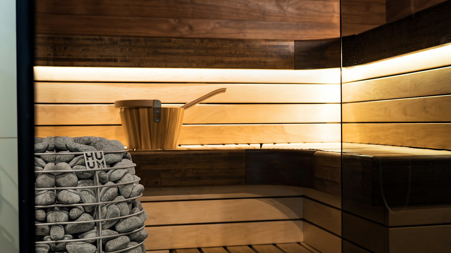 Wooden sauna interior with a stone heater, wooden bucket, and soft lighting beneath the benches.